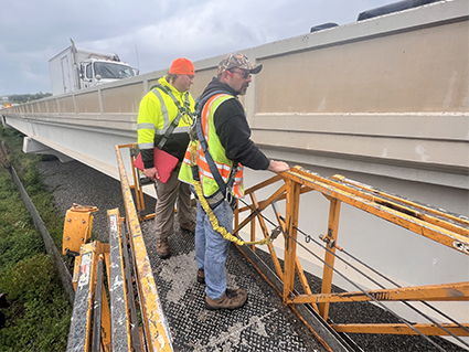 Two men in safety vests standing on a lift next to a bridge.