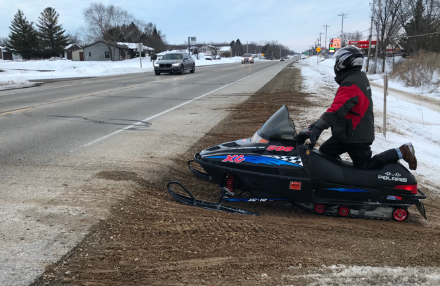A snowmobile at the side of highway with traffic, waiting for the right time to cross.