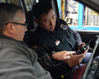 Trooper sitting in their patrol car with a person in the passenger seat looking at a network device.