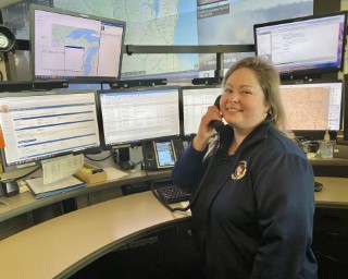 Dispatcher holding a phone to their ear turned and smiling at the camera while sitting at a desk with 6 monitor screens.