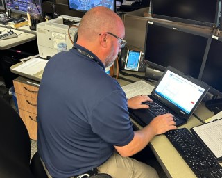 IT professional sitting at their desk working on a laptop with various other technology on the desk in the background.